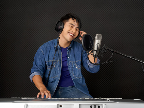 Young asian man with headphones playing an electric keyboard in front of black soundproofing walls. Musicians producing music in professional recording studio.