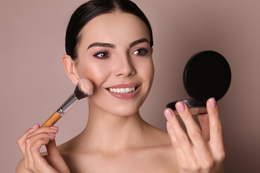 Beautiful young woman applying face powder with brush on dusty rose background, closeup