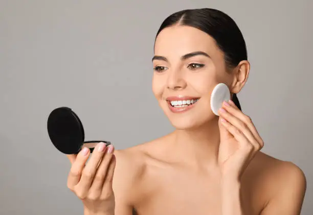 Beautiful young woman applying face powder with puff applicator on grey background