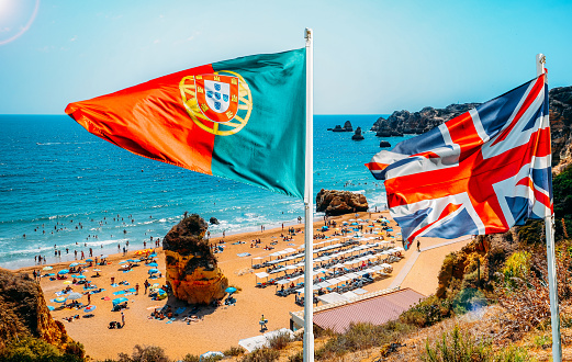 Portugal and United Kingdom flags on foreground with perspective view of beachgoers at Cova Redonda Beach in Algarve, southern Portugal on a summer day