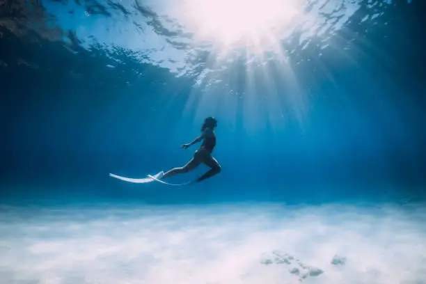 Freediver with white fins glides and posing underwater in ocean with sunlight.