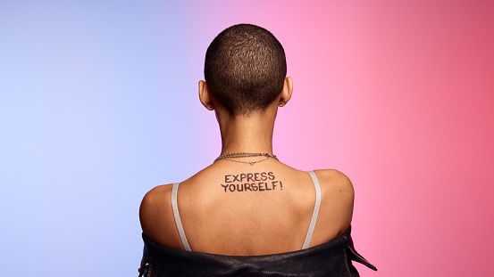 Rear view of unrecognizable female with express yourself written on her back. Female with shaved head on colorful background.