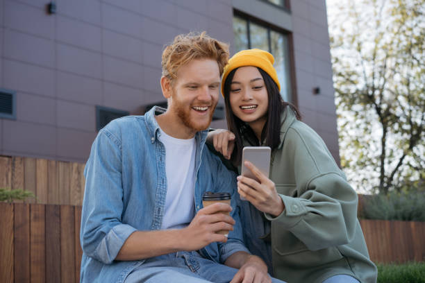 istock Young smiling friends using mobile phone watching videos, shopping online, communication together outdoors 1324510697