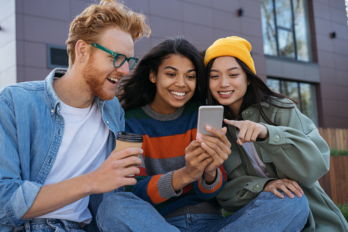 Group of smiling multiracial friends using mobile phone, watching video, shopping online, having fun outdoors. Young happy students studying, learning language, communication together