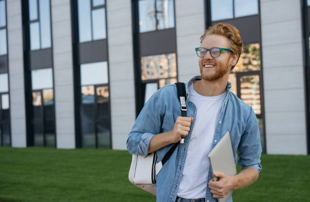 istock Confident man wearing eyeglasses looking away, copy space. Smiling university student with backpack and laptop computer walking in university campus, education concept 1324511040