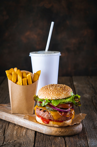 Burger combo with cheeseburger hamburger, take away beverage straw glass and disposable french fries in kraft paper box on dark wood table background