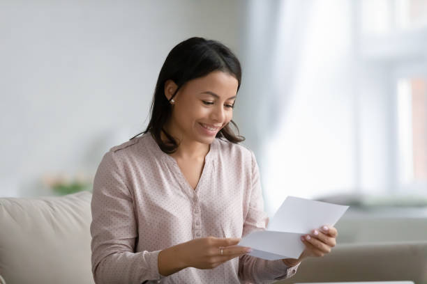 Happy young mixed race woman holding and reading paper letter Happy young mixed race woman holding and reading paper letter, receiving invitation or good news about approved loan, mortgage, tax, insurance, getting notice about acceptance of statement college acceptance letter stock pictures, royalty-free photos & images