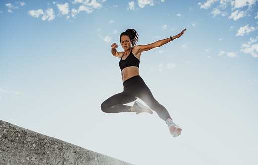 Fitness woman jumping from wall as a training routine. Low angle view of a female athlete in sportswear exercising outdoors in morning.
