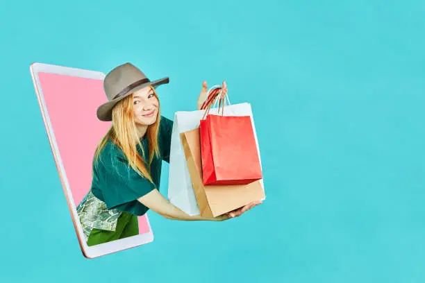 Photo of Virtual mobile buying. Young female holding shopping bag on online store or shop discount promotion look through the mobile or tablet screen. E-commerce, sale, discounts, advertising in app concept.