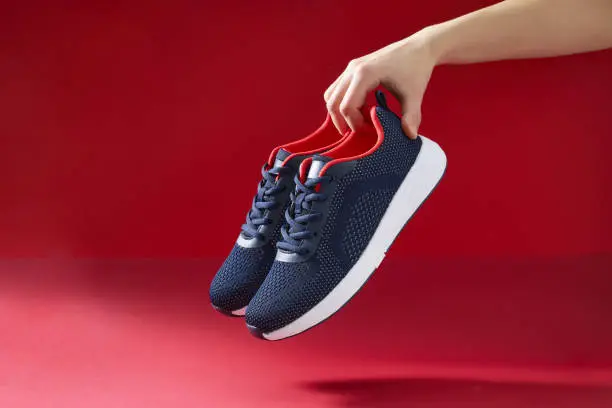 Photo of Hand hold sports shoes on red background. Holding new fashion sneakers for running. Choosing and buy of new sneakers.
