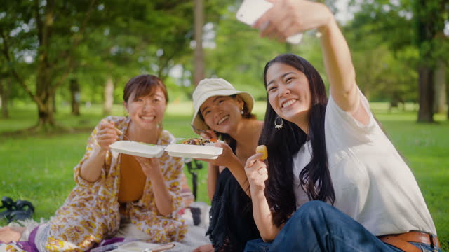 3人の女性の友人がピクニックを楽しみ、自然の中でスマートフォンでセルフィーを撮っています。