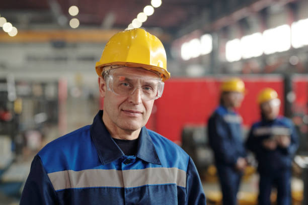 Portrait of content mature manufacturing worker Portrait of content handsome mature Caucasian manufacturing worker in work helmet and protective goggles standing against colleagues at factory manufacturing occupation stock pictures, royalty-free photos & images
