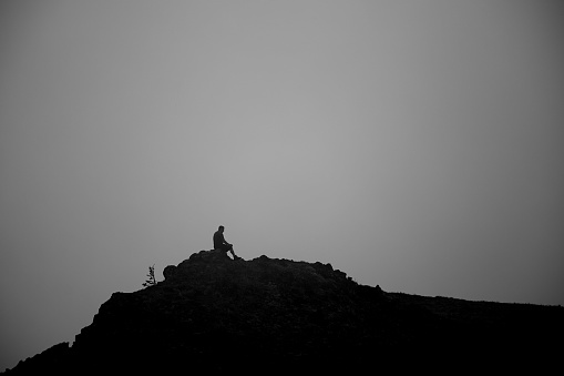 A lone hiker sits atop a rock outcropping in swirling mist in the Olympic Mountains of Washington State.