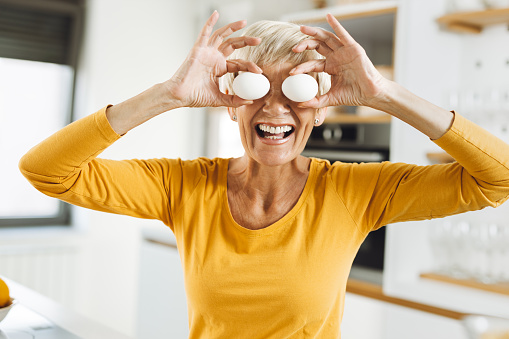 Happy senior woman covering her eyes with eggs and having fun in the kitchen