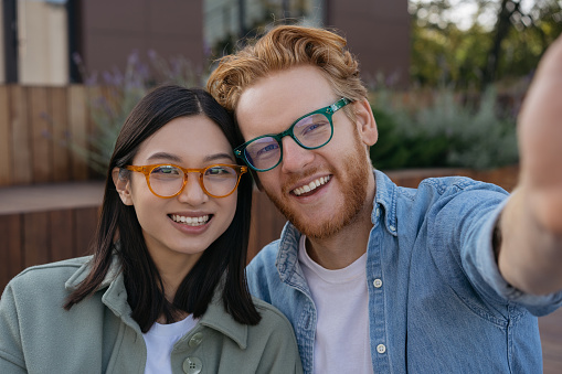 Young smiling friends wearing stylish eyeglasses taking selfie on the street. Portrait of beautiful asian woman and handsome caucasian man looking at camera. Positive lifestyle concept