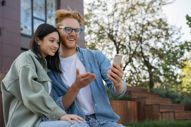 amici sorridenti che usano lo shopping del telefono cellulare online. bella donna asiatica e bella comunicazione uomo caucasico insieme, guardando video su smartphone seduti nel parco - smiling women glasses assistance foto e immagini stock