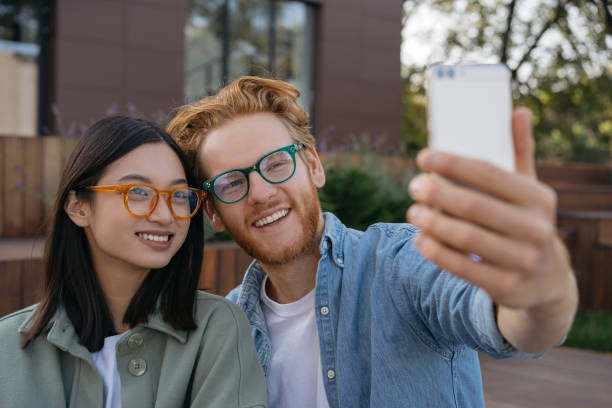 Smiling friends influencers using mobile phone, recording video outdoors. Young attractive asian woman and red haired caucasian man wearing stylish eyeglasses taking selfie on the street stock photo