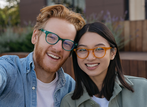 Close up portrait of smiling couple wearing stylish eyeglasses. Beautiful asian woman and handsome caucasian man embracing, looking at camera outdoors. Happy people in eyeglasses, vision concept