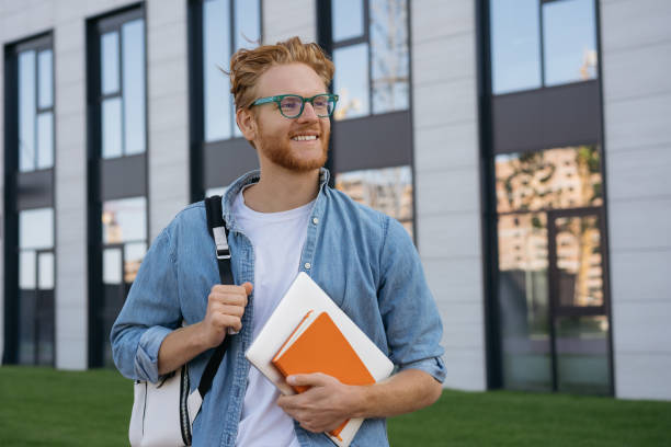 student z plecakiem i książki spaceru w kampusie uniwersyteckim, koncepcja edukacji. portret szczęśliwego przystojnego mężczyzny ubranego w stylowe okulary patrzące z dala od ulicy, kopiuj przestrzeń - men holding smiling young adult zdjęcia i obrazy z banku zdjęć