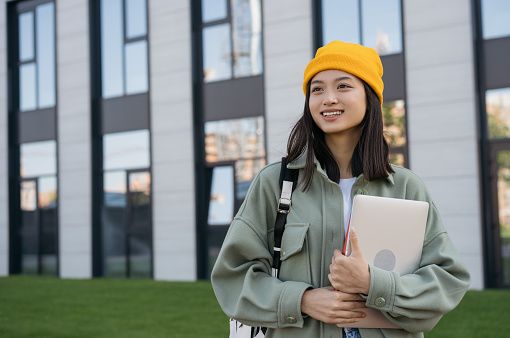 Portrait of smiling asian woman holding laptop computer looking away on the street. Happy student walking to university