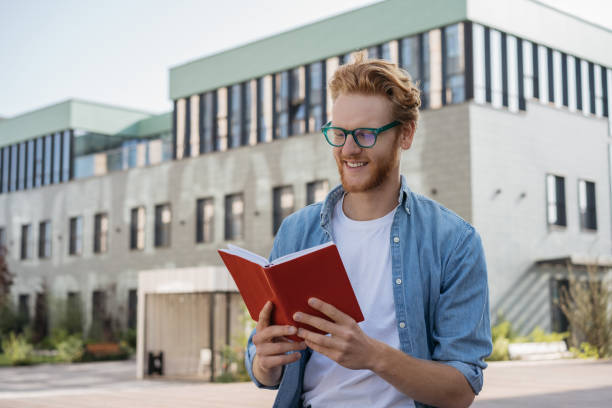 istock Smiling man reading book outdoors. Successful university student wearing stylish eyeglasses studying , exam preparation, education concept 1324585381