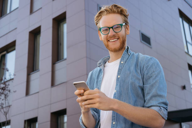 istock Smiling red haired man using mobile app for online shopping looking at camera, standing outdoors. Portrait of young successful freelancer wearing stylish eyeglasses receive payment 1324585449
