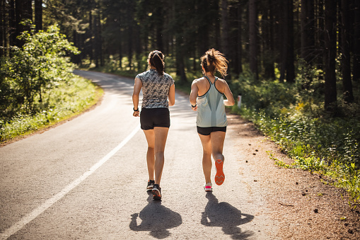 Back view of two female athletes running on road in forest on sunny day