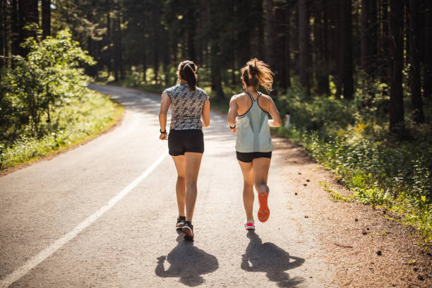 задний вид спортсменок, бегая по дороге в лесу - road running стоковые фото и изображения