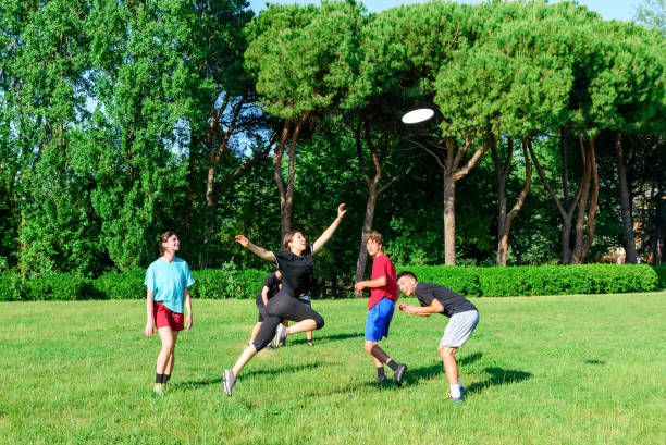 公園のアウドアでプラスチックフライングディスクゲームで遊ぶカジュアルウェアを着た混合若いティーンエイジャーの人々のグループ。ジャンプの女性は試合でチームメイトにディスクを� - men summer passing tossing ストックフォトと画像