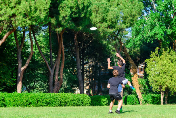 公園のアウドアでプラスチックフライングディスクゲームで遊ぶカジュアルウェアを着た混合若いティーンエイジャーの人々のグループ。ジャンプ男は試合でチームメイトにディスクをキャ� - men summer passing tossing ストックフォトと画像
