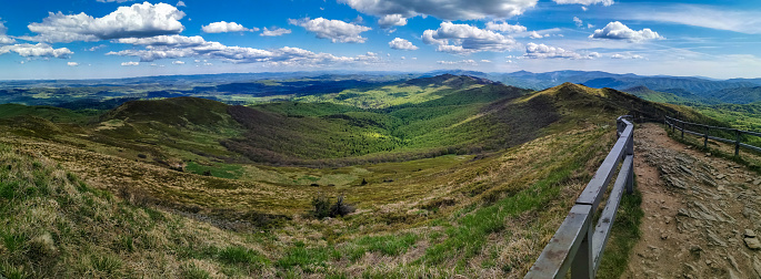 A postcard from the Bieszczady Mountains, a trail in the meadows and a view of the mountains, greenery, grass and trees and the blue sky. Rocky trail with railings