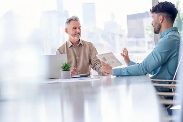 Businessmen working on financial data on a digital tablet. There are several graphs and charts visible on the digital tablet. It may be a training session. Both are dressed in business casual clothing in a board room. One man has grey hair