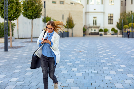 Young Urban Business Woman with a Smartphone in the Street is Heading Toward the Office. Staying in Touch with her Clients. Attractive Young Blonde in Business Suit and a Briefcase in the City Streets is in Rush Hour.