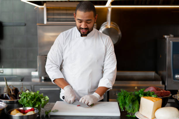 istock Handsome young African chef standing in professional kitchen in restaurant preparing a meal of meat and cheese vegetables. 1324612698