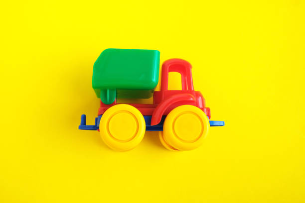 A close-up of a vibrant toy truck sitting in a sandy outdoor play area, partially surrounded by small leaves and grass