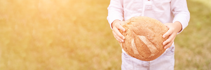 Child holds and bite round bread. Healthy food. Carrying big fresh baker bun. Rustic product. Enjoy warm breakfast. Craft cuisine. Home made lunch. Green outdoor background. Anonymous trend