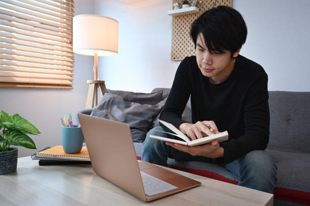 istock Young Asian man holding notebook and using laptop while sitting on couch. 1324635970