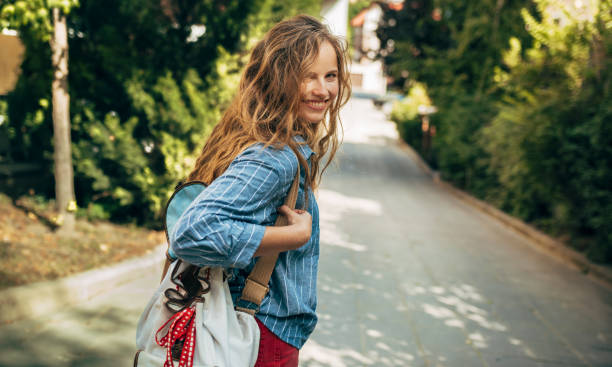 rearview of a beautiful young woman in a casual outfit and backpack posing outdoors. pretty female student with long red curly hair walking in the city street on a sunny day. - long hair red hair women men imagens e fotografias de stock
