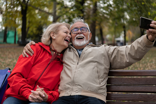 Portrait of Senior Couple Sitting on the Wooden Bench in Park After Shopping and Having a Lovely Conversation While Taking a Selfie Photos. A Couple of Retirees with Paper Bags is Relaxing in City Park.