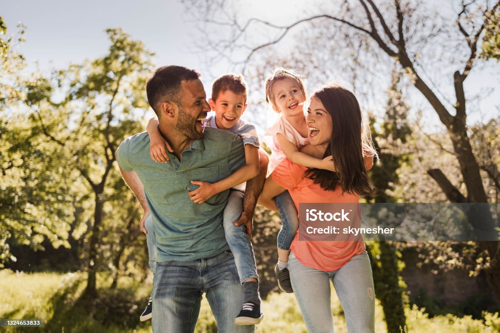 Happy parents having fun while piggybacking their small kids in nature. Playful family having fun while piggybacking in springtime. Family Stock Photo