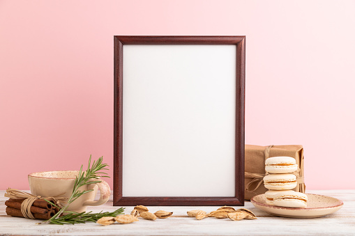 Brown wooden frame mockup with cup of coffee, almonds and macaroons on pink pastel background. Blank, side view, still life.