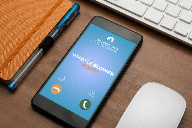 istock Whistleblowing concept: smartphone next to an orange notepad on wooden table show a whistle blower alert on an anonymous call 1324665061