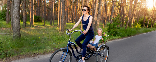 Young adult caucasian mom enjoy having leisure fun riding bicycle with cute adorable blond daughter holding wild field flower at scenic rural country road on bright sunny day. Countryside vacation.