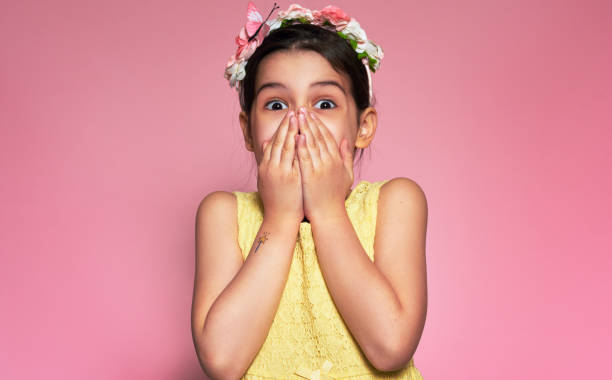 Candid portrait of an astonished little girl wearing a yellow dress covering her mouth with hands receiving a gift at her birthday. Kid covering lips with hands for the mistake on pink background stock photo
