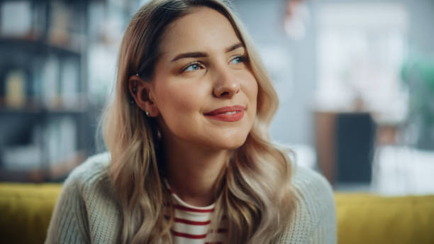 portrait de belle jeune femme aux cheveux brun clair portant un pull en laine blanche regardant vers le haut et souriant avec charme. femme réussie se reposant dans un salon lumineux. - charmingly photos et images de collection