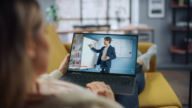 istock Beautiful Caucasian Female is Using Laptop Computer for Watching Online Video Lecture. She is Lying on a Couch Sofa at Home Living Room. University Student Studies From Home during Pandemic. 1324677811