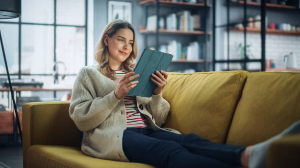 istock Beautiful Caucasian Female Using Tablet Computer in Stylish Living Room while Resting on a Cozy Couch Sofa. Young Woman at Home, Browsing Internet, Using Social Networks, Having Fun in Flat. 1324677836