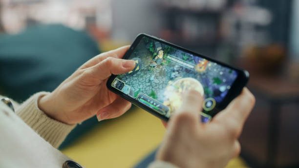 gros plan sur les mains jouer à un jeu vidéo de stratégie rpg sur un smartphone tenu horizontalement à la maison salon. mains féminines tapant sur l’écran du téléphone mobile avec un jeu coloré sur internet. - geek application photos et images de collection