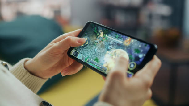 close up on hands playing an rpg strategy video game on a horizontally held smartphone at home living room. feminine hands tapping the mobile phone screen with a colorful game over the internet. - equipamento de jogo imagens e fotografias de stock