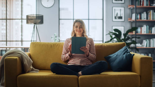 istock Beautiful Caucasian Female Using Tablet Computer in Stylish Living Room while Sitting on a Cozy Couch Sofa. Young Woman Resting at Home, Browsing Internet, Using Social Networks, Having Fun in Flat. 1324678080